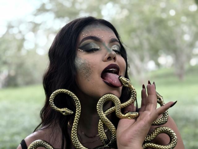 Lara James, com cabelo escuro, sombra verde e batom preto, língua de fora, segurando serpentes douradas no rosto e na mão, com unhas longas, com uma aparência sexy e selvagem..