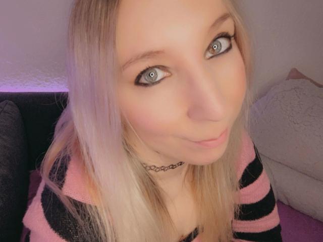 JustKerry-hot with blue eyes, blonde hair, pink and black striped shirt, choker, and a cute smile, showing off her tits slightly. Soft pink and white background.