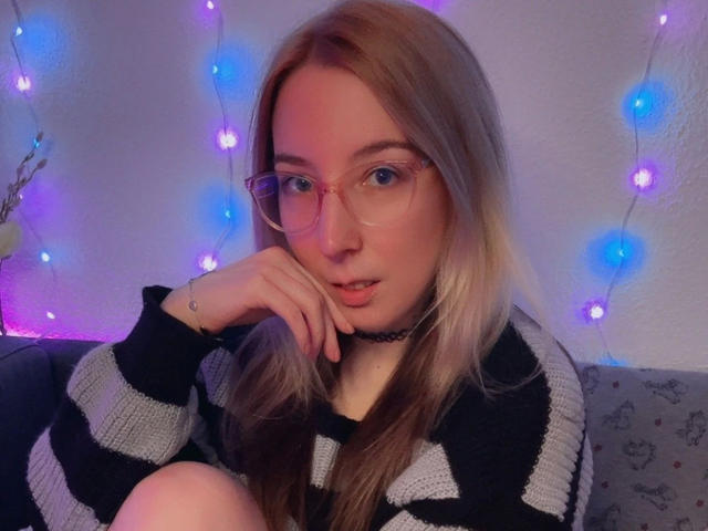 JustKerry-hot with glasses, blonde hair, black-and-gray striped sweater, choker, and blue-purple fairy lights in the background, looking cute and slightly bored.