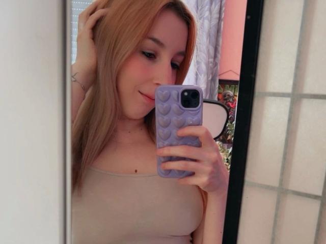 JustKerry-hot with blonde hair, holding a purple phone with heart patterns, taking a mirror selfie, wearing a beige top, smiling slightly, hand in hair, visible star tattoo.