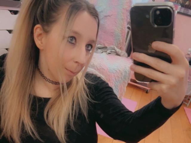 JustKerry-hot taking a selfie, blonde in a ponytail, black long-sleeve shirt, choker, holding a phone, blue eyes, wooden floor, mirror reflection, slightly smiling, cute.