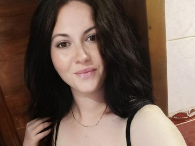 MaluskaHot with black hair, light skin, brown eyes, and pink lips, wearing a black top, smiling slightly, and touching her hair with a gold necklace.