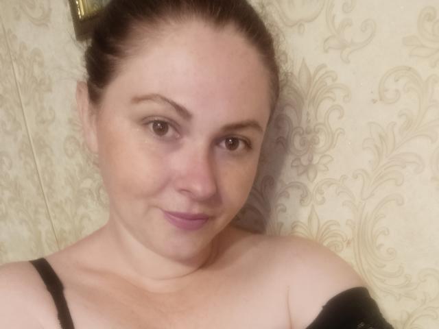 MaluskaHot with light skin, brown eyes, and brown hair in a bun, smiling with pink lips, wearing a black strap, has a cute, chubby face and small tits.