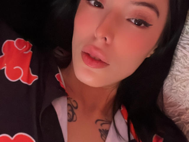 AizenMyoo with black hair, pink lips, and tattoos, wearing a black robe with red clouds, has winged eyeliner, lying on a white textured surface, looking up.