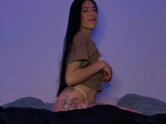 AizenMyoo met lang zwart haar, een bruin shirt, een witte string en een tatoeage op haar rechter bil, lachend naar de camera, zittend op een bed met grijze kussens..