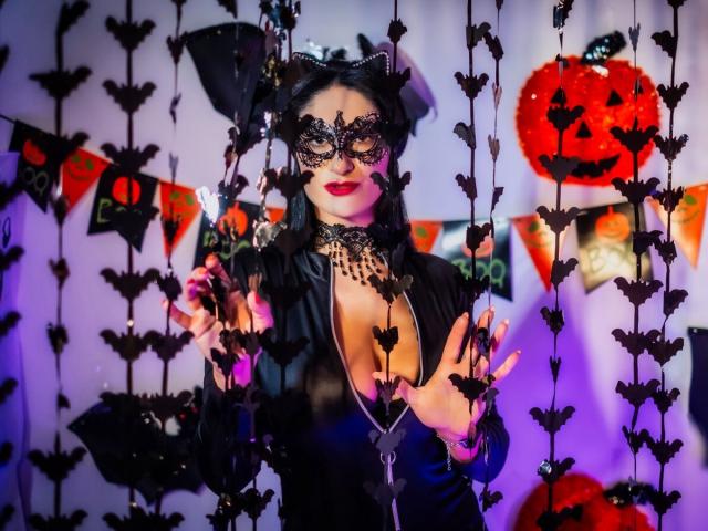 HotSweetPie con máscara de gato negro, collar de encaje y escote pronunciado, vestida con un conjunto de cuero negro, sosteniendo cuerdas con forma de murciélago, faroles rojos y decoraciones de Halloween en una habitación iluminada con luz púrpura..