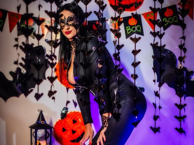 HotSweetPie, atuendo de cuero negro, escote pronunciado, máscara de encaje negro, pantalones negros, cabello negro, lápiz labial rojo, inclinada hacia adelante, decoraciones de Halloween, murciélagos, farol, iluminación púrpura y azul, curvilínea..