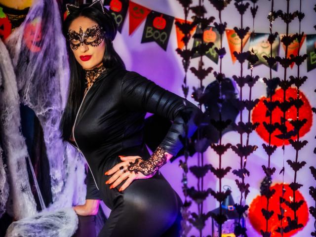 HotSweetPie con orejas de gato negras, una máscara de encaje y un conjunto de cuero negro, mostrando su curvilínea figura y sus manos cubiertas de encaje, de pie junto a una telaraña..
