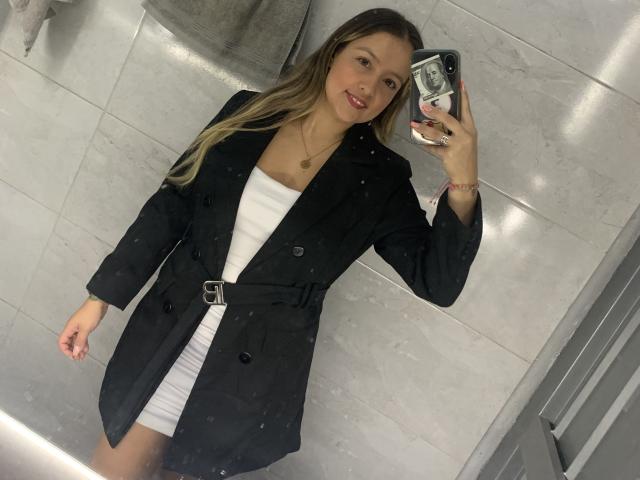 SaritaCaramelo with blonde hair, black coat, white dress, holding phone, smiling, mirror selfie, visible tits, pink lipstick, gray towel, marble tiles, ring on neck.