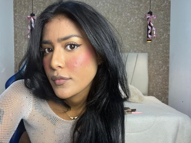 Babilett heeft lang zwart haar, bruine ogen met een vleugje eyeliner, roze blush en draagt een witte top van gaas. Ze kijkt serieus in de camera..