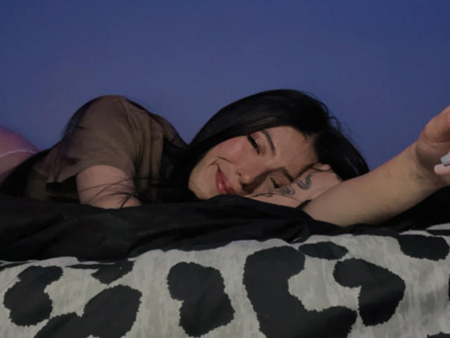 AizenMyoo with black hair, lying on a bed, eyes closed, smiling, arm raised, wearing a sheer brown top, tattoo on right shoulder, black and white patterned blanket.