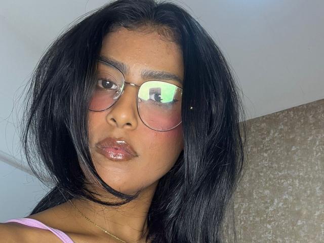 Babilett has dark black hair, wearing reflective glasses, pinkish lipstick, and a pink top, with a subtle gold necklace, looking serious. Her face shows slight redness around the eyes.