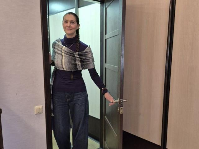 TayaFlavy with medium tits, wearing a plaid shawl over a navy turtleneck, blue jeans, and a braid, standing in a doorway with a slight smile.