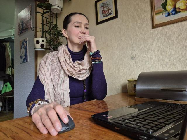 TayaFlavy, with a scarf, dark top, and bracelets, sits at a wooden table, thinking, with a hand on her chin, and her other hand on a black laptop.