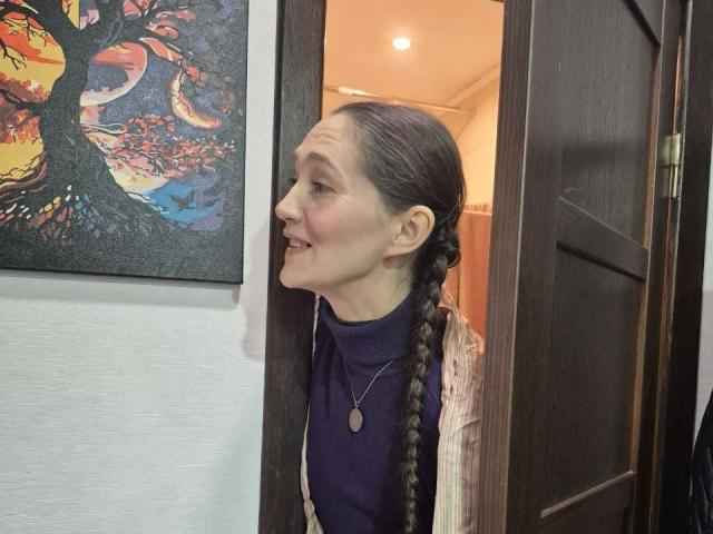 TayaFlavy with a smile, brown braid, navy shirt, pink cardigan, and necklace, standing in a dark wooden door frame, looking to the left, with a colorful painting on the wall.