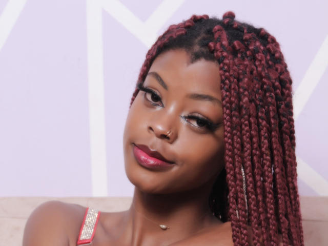 AlixNoa with dark red braided hair, shiny brown skin, pink lipstick, nose ring, and a slight smile. She's wearing a pink and gold top.