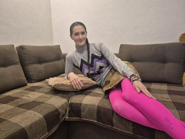 TayaFlavy con medias rosas, camiseta gris con estampado de zigzag, falda marrón, sofá marrón a cuadros, acostada sobre un cojín, sonriendo, cabello oscuro en trenza, múltiples pulseras, mano derecha sobre el muslo, mano izquierda sobre el cojín..