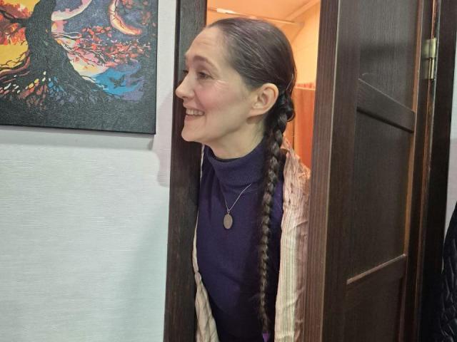TayaFlavy with braided brown hair, smiling, wearing a purple shirt and checkered cardigan, standing in a doorway, wearing a necklace, looking to the left.