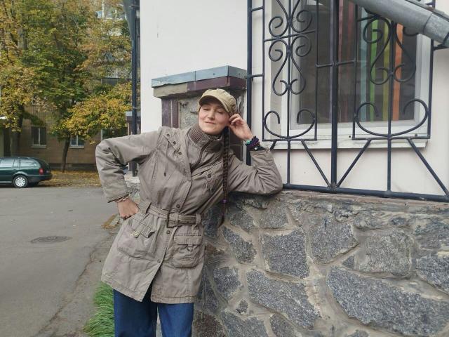 TayaFlavy leans against a stone wall in a beige trench coat, blue jeans, and a beige cap, hand on hip, smiling with a slight touch to her cap.
