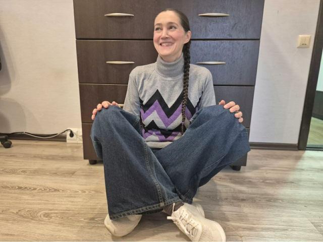 TayaFlavy assise sur un plancher en bois, les jambes relevées, portant un pull gris à motifs en zigzag, un jean bleu, des baskets blanches, souriante, avec de longs cheveux tressés, une commode marron derrière elle, les mains posées sur les genoux..