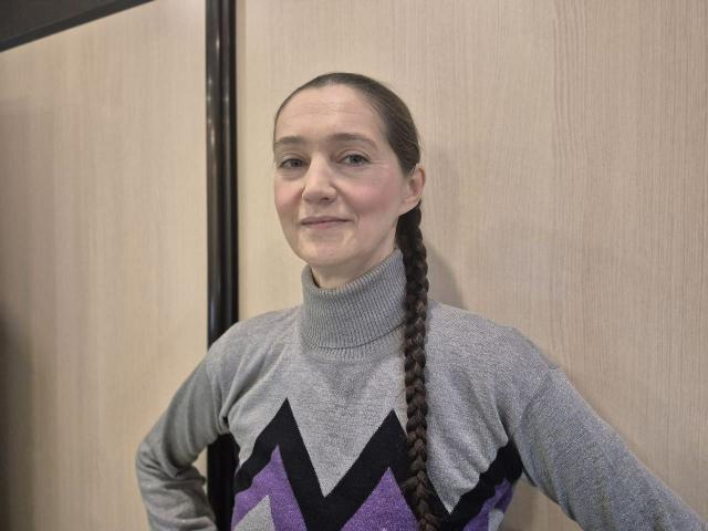 TayaFlavy with a light gray, patterned sweater, purple and black geometric design, and a long braid, smiling, standing against a wooden wall.