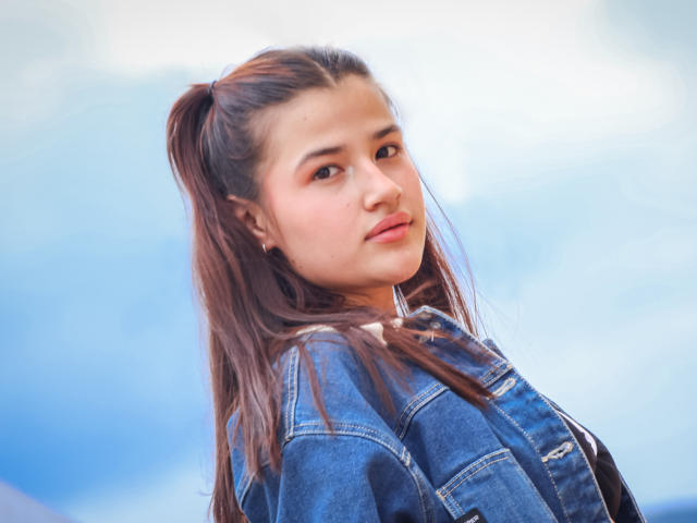 IrisBlair with long brown hair in a ponytail, wearing a blue denim jacket, looking over her shoulder with a slight smile, soft blue sky in the background.