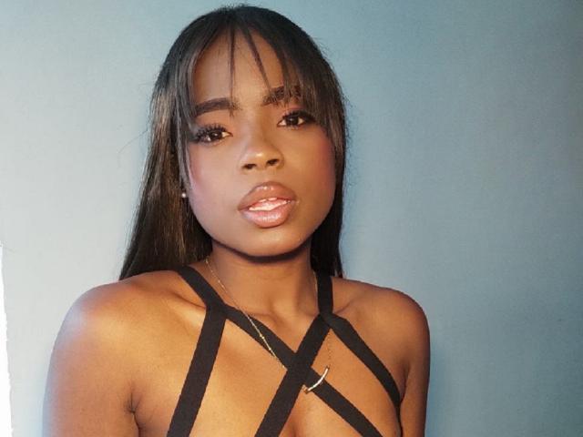 LindaBird with dark brown skin, long straight black hair, and bangs, wearing a black strappy top, showing her shiny pink lips and small gold necklace.
