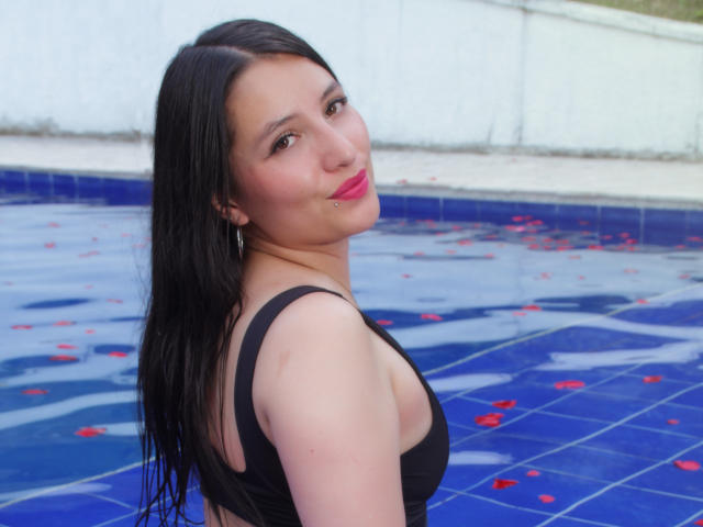 SamyNoa with wet black hair, wearing a black tank top, by a blue-tiled pool with red rose petals, smiling with pink lipstick, looking over her shoulder.