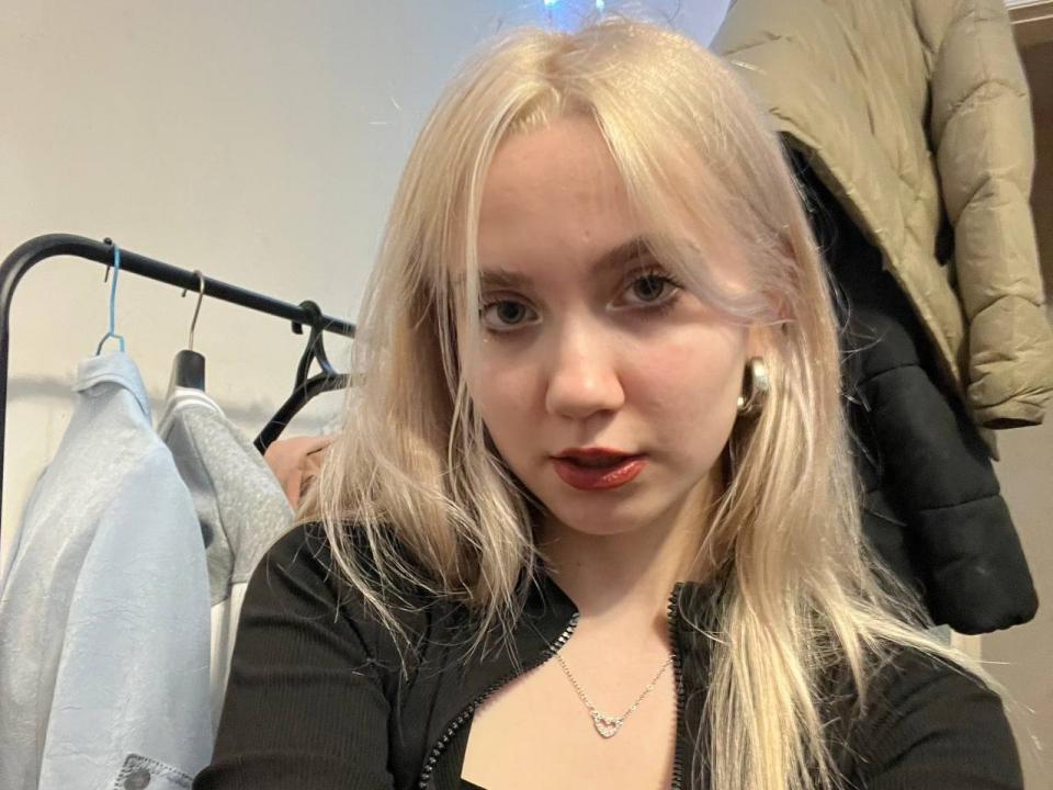 ReginaPatty with blonde hair, red lipstick, black zipped-up top, small necklace, and white earbuds, looking at camera, clothes hanging in background.