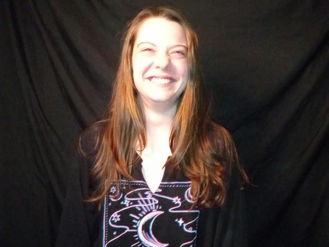 GoddessHott69 has long brown hair, a white skin tone, and a smiling face. She's wearing a black shirt with a white, colorful, crescent moon design.