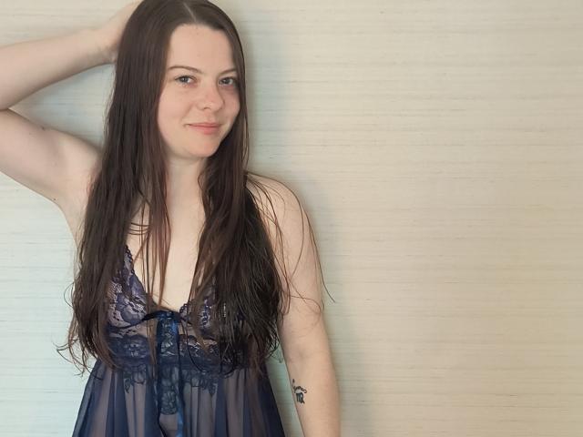 GoddessHott69 with long brown hair, wearing a sheer blue lace nightie, exposing her small tits, standing against a plain wall, hand in hair, subtle smile.