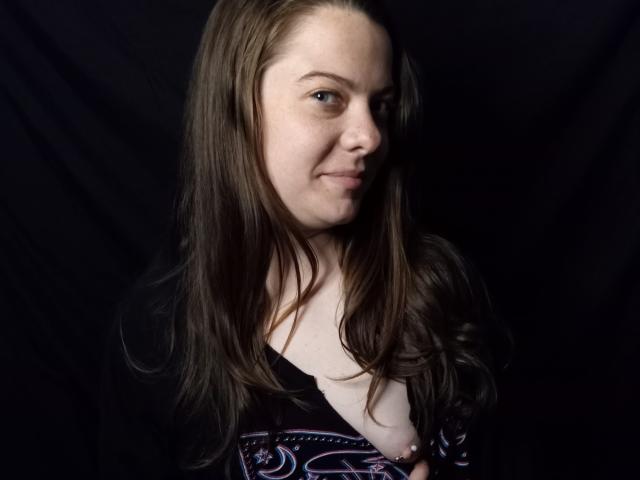 GoddessHott69 with long brown hair, fair skin, and a subtle smile, wearing a black patterned top, showing a small pink nipple, against a dark background.