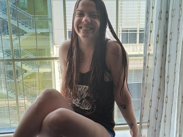 GoddessHott69 with long brown hair, smiling, wearing a black tank top, and blue shorts, sitting by a window, showing her ass and a tattoo on her right arm.