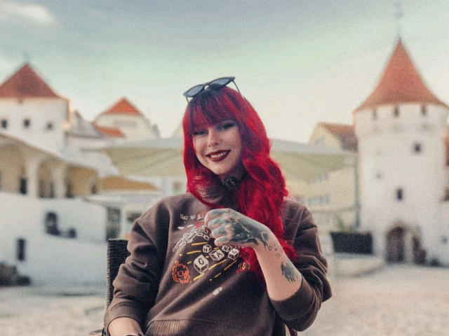 Teufelshelferin-hot with red hair, wearing a brown sweater, showing a tattooed hand, smiling with red lipstick, sunglasses on head, standing in a cobblestone courtyard with white towers.