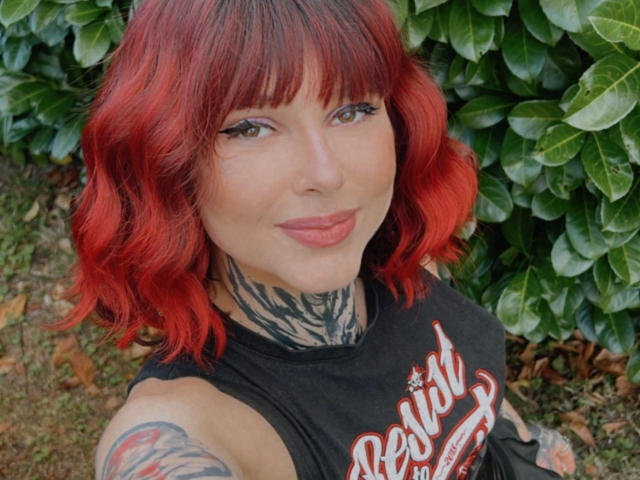 Teufelshelferin-hot has red hair, tattoos on neck and arm, and is wearing a black sleeveless shirt, showing a cute smile with pink lips, standing outside.