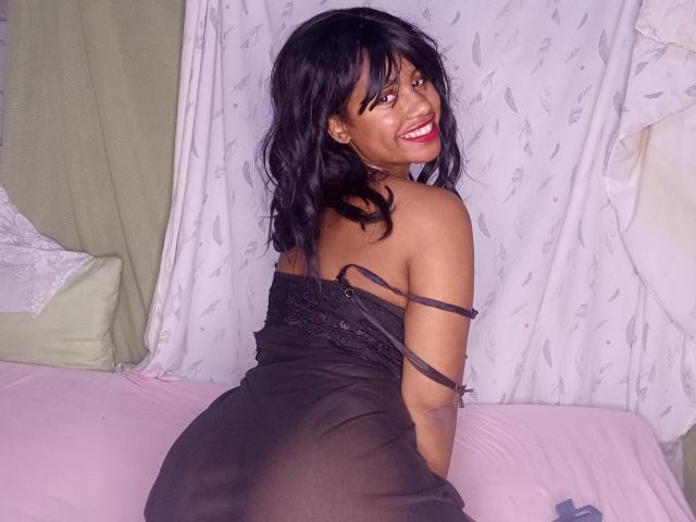 LayahCoquine with black hair, smiling, wearing a sheer black dress, showing her ass, sitting on a pink bed, green and white curtains in the background.