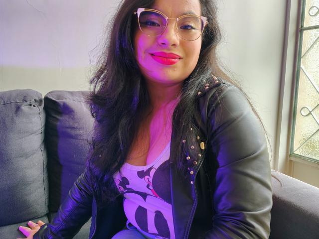 MaayGrey with long black hair, glasses, and pink lipstick, wearing a black leather jacket with studs over a Minnie Mouse shirt, sitting on a gray couch.