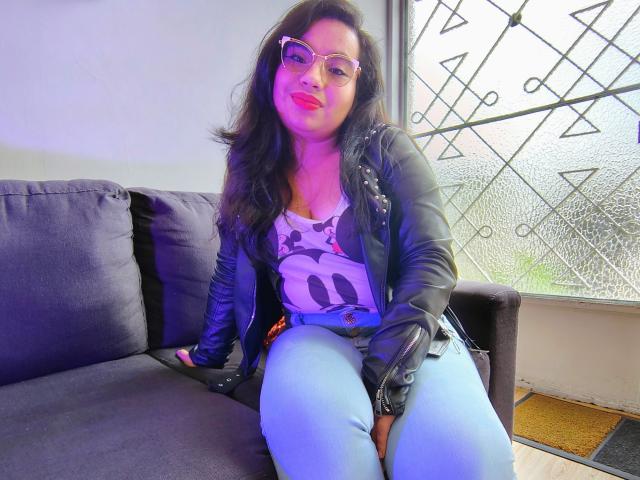 MaayGrey with glasses, pink lipstick, black leather jacket, white tank top, blue jeans, sitting on dark sofa, window with geometric pattern in background. Boobs visible under tank top.