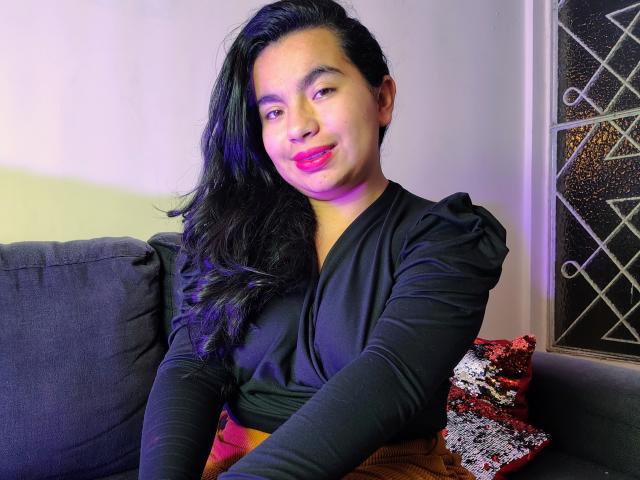 MaayGrey has long black hair, pink lipstick, and a black V-neck top. She's sitting on a grey couch with a patterned pillow, smiling slightly. Her ass is hidden.