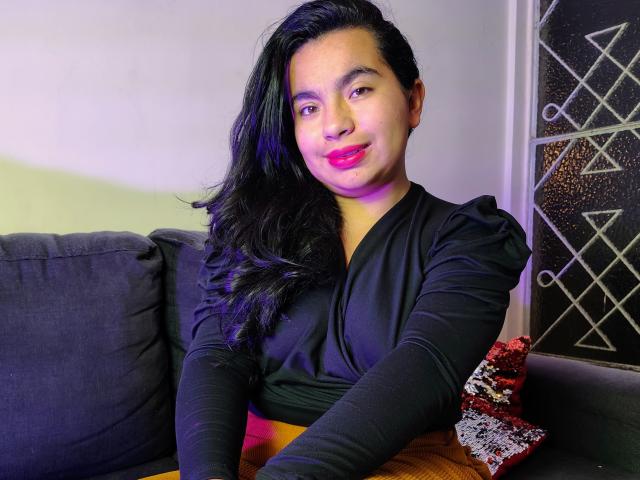 MaayGrey with long black hair, pink lipstick, and black dress, sitting on a gray couch, smiling slightly, with a decorative pillow on the right.