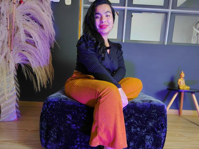 MaayGrey, sitting on a black velvet stool, wearing a black long-sleeve shirt and orange pants, with black hair, red lipstick, and a slight smile, in a colorful room.