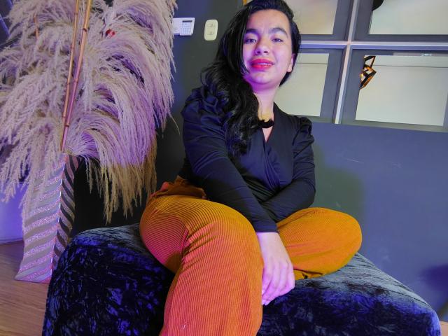 MaayGrey with black hair, red lipstick, black top, and orange ribbed pants sits on a velvet chair, legs crossed, hands on knees, puffy ass, calm expression.