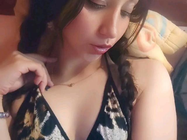 GhanimaY with braided brown hair, wearing a leopard print halter top, has a hand on her chest, showing her small tits and a gold necklace, with a slightly open mouth.