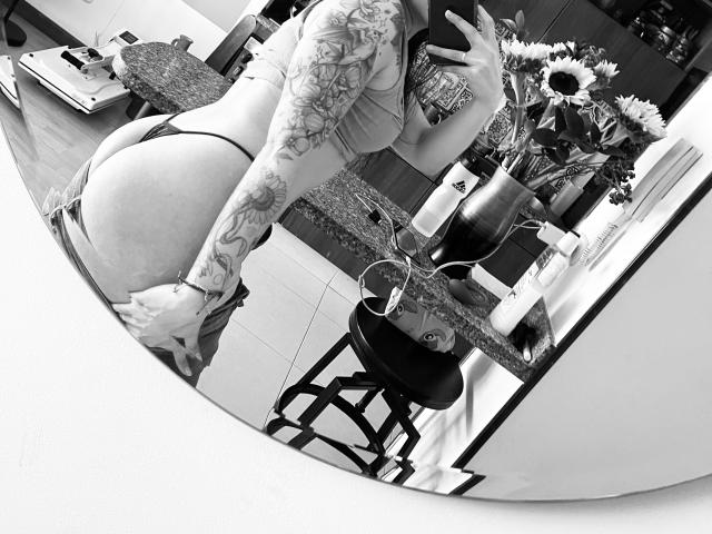 Black-and-white mirror selfie of SujannaVarma with tattooed arm, pulling down thong to reveal her big ass. She's wearing a bra and holding her phone. Sunflowers in the background.