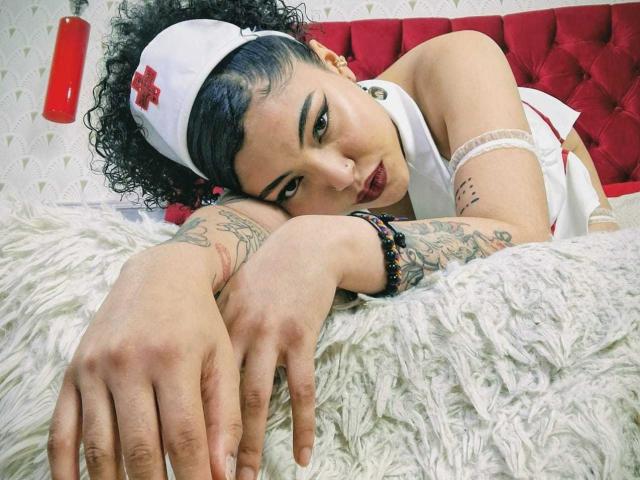 SammyHills69 mit lockigem, schwarzem Haar, weißer Krankenschwesteruniform, rotem Kreuz-Haarband, Tattoos, dunklem Lippenstift, einer weißen Felldecke, einem roten, gepolsterten Kopfteil und einem schwarzen Armband, liegt im Bett und blickt nach unten..
