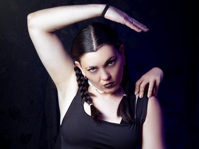 SaharaMK with dark braided hair, black sleeveless top, arm raised, looking intense, black lipstick, white ear clip, dark background, blue light, right arm on shoulder, black wristband.
