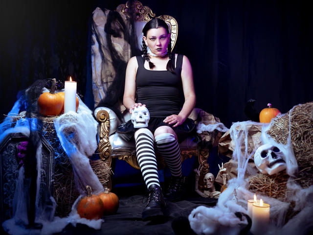 SaharaMK, with black tank top, striped thigh-highs, and boots, sits on ornate chair, holding white mask, surrounded by candles, pumpkins, skeletons, and spooky decorations.