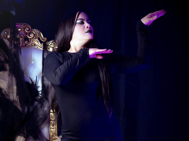 SaharaMK avec un maquillage sombre, de longs cheveux noirs, une robe noire, les yeux fermés, les bras levés, un éclairage violet, un fauteuil doré orné, un miroir reflétant ses fesses, et des ombres de paumes violettes-roses..