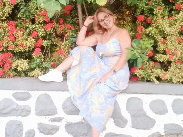 JaneTurner, middle-aged with glasses, sitting on stone wall, wearing floral dress, white socks, smiling, right hand touching head, left leg raised, red flowers and green leaves in background.