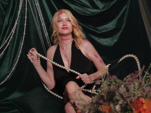 VelvetSkyy with blonde hair, red lipstick, black halter dress, holding pearl necklace, tattoos on arm, sitting against dark green velvet, bouquet in foreground, red marks on face and arms.